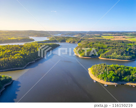 Aerial view of Orlik Water Reservoir on the Vltava River in Czechia, surrounded by forests and hills. Calm waters reflect blue sky, creating serene and idyllic landscape. Aerial view of Orlik Water Reservoir on the Vltava River in Czechia, surrounded by forests and hills. Calm waters reflect blue sky, creating serene and idyllic landscape. 116150219