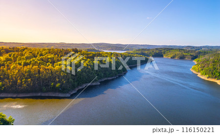 Aerial view of Orlik Water Reservoir on the Vltava River in Czechia, surrounded by forests and hills. Calm waters reflect blue sky, creating serene and idyllic landscape. 116150221