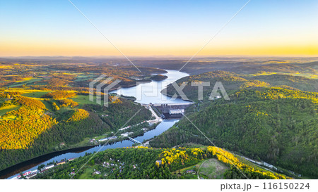 A sunset aerial shot of Orlik Reservoir in Czechia shows a winding river, hills, and a quaint town in the valley with vibrant blue and golden skies. 116150224