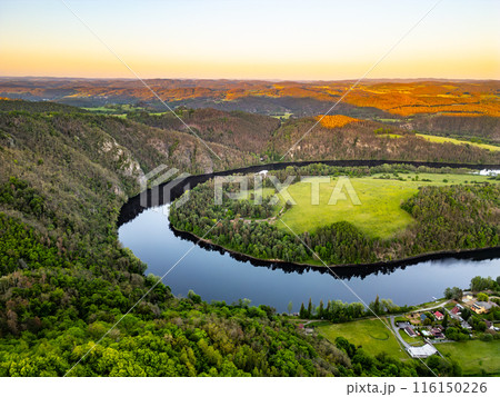 A scenic aerial view of Vltava River bending, Solenice Horseshoe, through the Czech countryside at sunset. Lush green trees and fields around river as the sun casts a golden glow over the landscape. 116150226