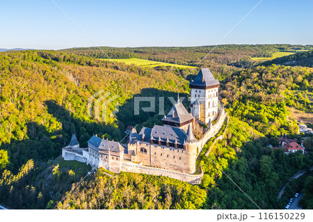 Aerial perspective of Karlstejn Castle nestled in a forested landscape with surrounding lush greenery on a clear, sunny day in Czechia. Aerial view from drone. 116150229