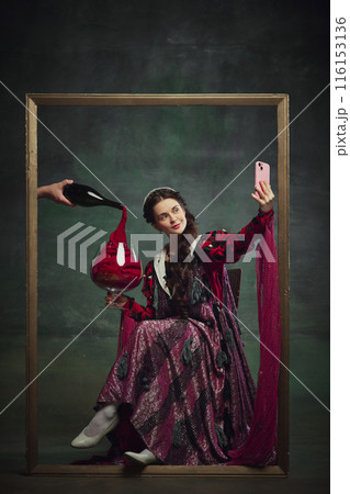 Portrait of young medieval royal person, woman sitting inside golden frame, with giant wine glass and taking selfie with mobile phone against vintage green background. Portrait of young medieval royal person, woman sitting inside golden frame, with giant wine glass and taking selfie with mobile phone against vintage green background. 116153136