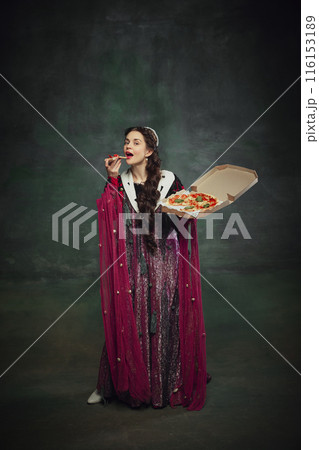 Humorous ads for pizza brands. Portrait of young woman, royal person in rich dress, eating pizza against vintage green background. Humorous ads for pizza brands. Portrait of young woman, royal person in rich dress, eating pizza against vintage green background. 116153189