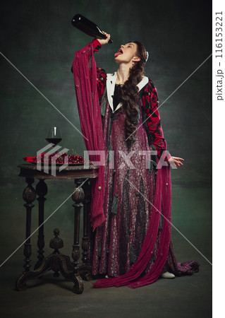 Addiction to alcohol. Young elegant woman, royal person shaking empty wine bottle on her mouth against dark green background. Addiction to alcohol. Young elegant woman, royal person shaking empty wine bottle on her mouth against dark green background. 116153221