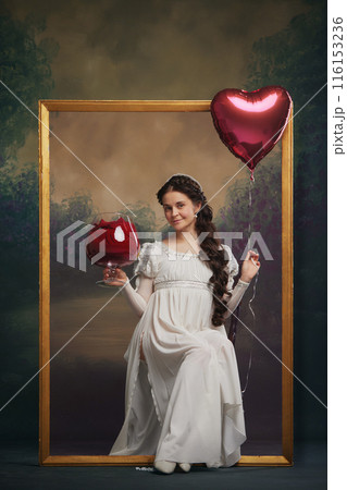 Portrait of beautiful young woman in tender white dress, sitting inside golden picture frame with heart-shaped balloon and giant wine glass. Valentine's day 116153236