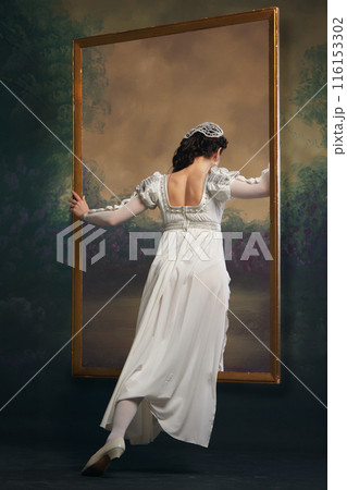 Looking into past. Young medieval woman in white dress looking inside picture frame against vintage background. Window to another era Looking into past. Young medieval woman in white dress looking inside picture frame against vintage background. Window to another era 116153302