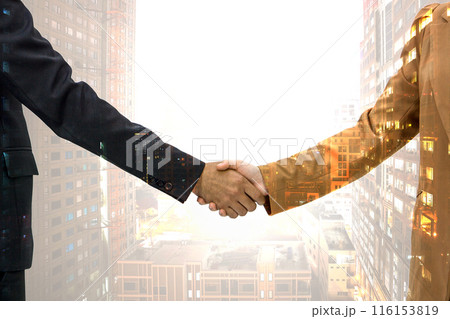 Two business people in suit are shaking hand, symbolizing agreement and partnership, with high rise office building cityscape overlay in the background. 116153819