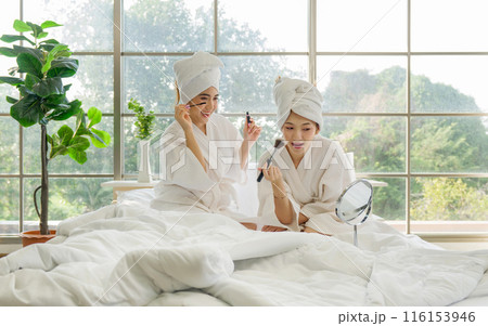 Two asian women are sitting relax on a bed in white robe and towel wrapped around their head, doing makeup with a mirror. 116153946