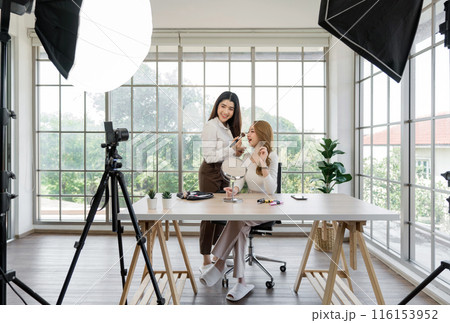 Two asian women creating content for social media. They are using a studio setup that include a camera mounted on a tripod, lighting equipment and various makeup product on the table. Two asian women creating content for social media. They are using a studio setup that include a camera mounted on a tripod, lighting equipment and various makeup product on the table. 116153952