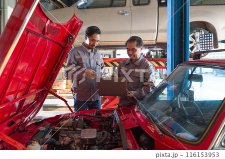 Two mechanic in gray uniform maintenance or inspection work on a vehicle while holding laptop computer. Diagnostic, referencing repair manual, or updating service record. Two mechanic in gray uniform maintenance or inspection work on a vehicle while holding laptop computer. Diagnostic, referencing repair manual, or updating service record. 116153953