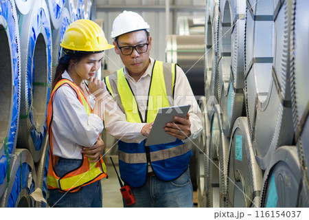 Senior engineer manager trains new employee within the metal sheet factory. Everyone wear safety vest and hardhat. Steel sheet roll stack are in the working area. 116154037