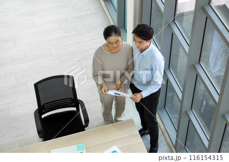 Two people in an office look at a paper with graphs. They stand next to a large window discussing it. 116154315