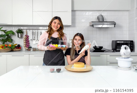 Two young women in apron make dessert in the kitchen. One people using a piping bag, pipe the icing onto the cake. The other holding wooden tray with a handful of colorful baking tools or decoration. 116154395