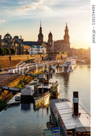 Scenic warm sunset beautiful Dresden city skyline at Elbe River with many steam tourist ship vessel moored at bank. Saxony Germany capital cityscape dusk vertical view with old Europe architecture 116154495