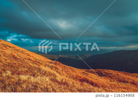 A scenic landscape view of a mountain range with a dramatic sky. The image features a foreground of rolling hills with dry grass and a distant mountain range covered in clouds. The sky is a vibrant 116154908
