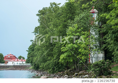 old lighthouse on the seashore among the trees against the backdrop of ancient buildings and embankment 116156730