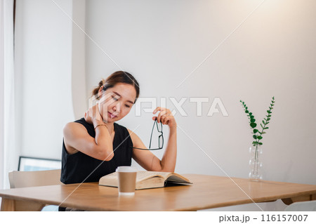 A young woman sits at a wooden table reading a book, holding her glasses, with a coffee cup and a plant in a vase nearby in a minimalist, bright room. 116157007