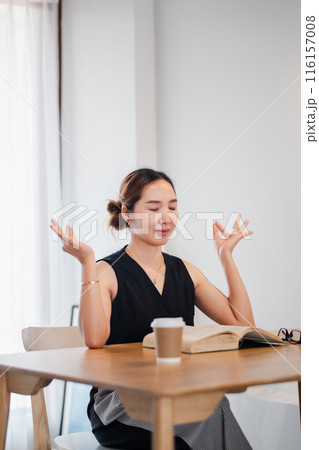 A woman sits at a wooden table with a book and coffee, practicing mindfulness meditation in a bright, serene room. A woman sits at a wooden table with a book and coffee, practicing mindfulness meditation in a bright, serene room. 116157008