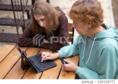 computer awareness of the elderly. a happy teenage girl teaches older 70s grandmothers to use a laptop at home. social media and modern technologies 116158771