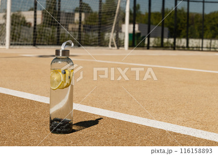 Transparent refreshing glass bottle of water in sunny day with hard shadow on outdoor soccer sports ground. Concept water balance or isotonic for workout. Copy space. 116158893