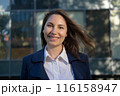 Portrait of an attractive young adult woman in a blue suit standing against the background of an office building, smiling and looking at the camera close-up 116158947