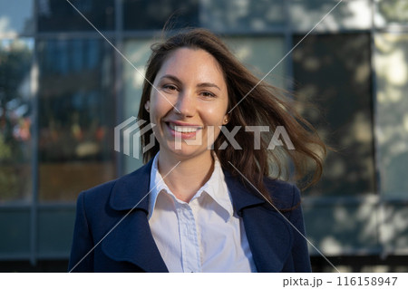 Portrait of an attractive young adult woman in a blue suit standing against the background of an office building, smiling and looking at the camera close-up Portrait of an attractive young adult woman in a blue suit standing against the background of an office building, smiling and looking at the camera close-up 116158947