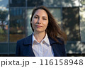 Portrait of an attractive strong confident independent woman in a blue suit standing against the background of a business building and looking at the camera 116158948