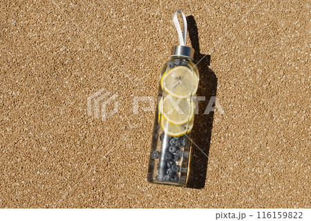 Transparent glass bottle of refreshing water with lemon and blueberries in sunny day on outdoor beige sports ground. Concept water balance for heat season for workout. View from above. Copy space. 116159822