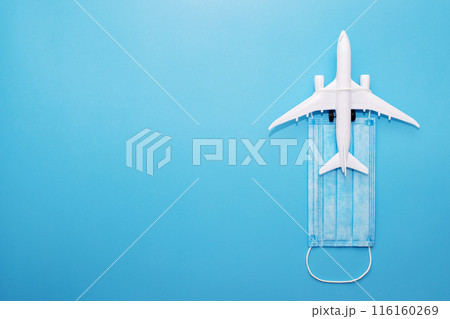 A plane model with a medical mask on a blue background. Safety flight and travel during quarantine and lockdown. Safe travels concept. Opening borders 116160269