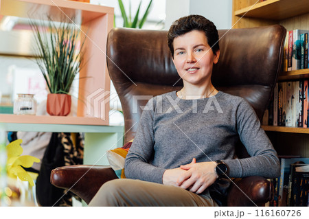 Confident, relaxed middle aged female expert sits in a leather armchair in cozy office with bookshelf background. 40s middle-age professional portrait of teacher, coach, mentor, therapist, counselor. Confident, relaxed middle aged female expert sits in a leather armchair in cozy office with bookshelf background. 40s middle-age professional portrait of teacher, coach, mentor, therapist, counselor. 116160726