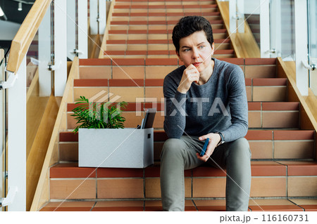 Stressed worried middle aged gender neutral woman with box of personal items sitting alone on the staircase after being fired, lost job due to recession and economic stress. Equality discrimination. 116160731