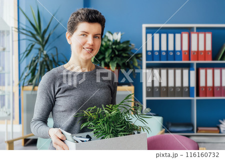 Portrait of smiling middle aged gender neutral woman hired by company holding box looking at camera on first day at work, happy new employee standing on new workplace in office with belongings. Portrait of smiling middle aged gender neutral woman hired by company holding box looking at camera on first day at work, happy new employee standing on new workplace in office with belongings. 116160732