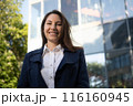 Portrait of a happy caucasian brunette woman in a blue suit stands against the background of a business building in the park and smiles 116160945