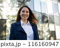 Portrait of a happy caucasian brunette woman in a blue suit stands against the background of a business building and smiles 116160947