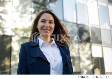 Portrait of a happy caucasian brunette woman in a blue suit stands against the background of a business building and smiles 116160947
