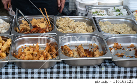 Assorted Asian street food in stainless steel trays, fried squid, various dumplings, spring rolls at food market 116162123
