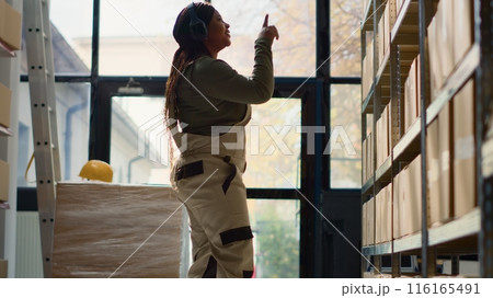 Happy warehouse manager listening music enjoying herself while examining merchandise. Cheerful supervisor wearing heaphones and dancing around while walking through storage facility rows 116165491