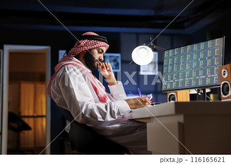 Middle Eastern guy sat at his workstation, intently researching and taking notes from his desktop computer. This image displays an Arab man with a notepad who is brainstorming and using a computer. 116165621