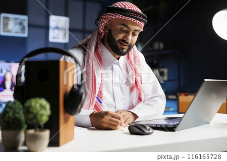 An engrossed male entrepreneur, dressed professionally, sits at a desk with his personal computer. Muslim man surfs the net on his laptop, surrounded by a notepad and focused on his online research. 116165728