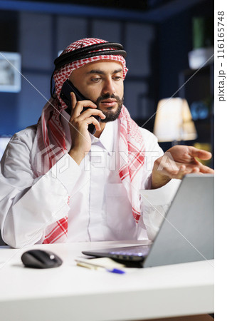 Detailed shot of a Muslim man concentrating on a phone conversation while checking his laptop. Arab guy who embraces technology demonstrates professionalism and strong communication skills. 116165748