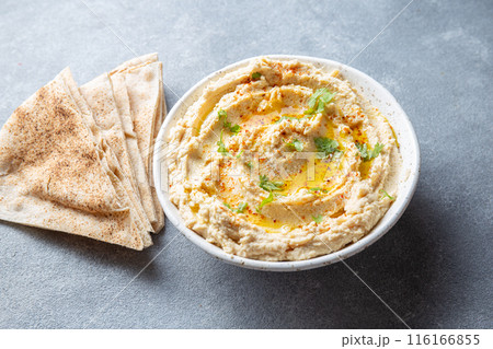 Bowl of creamy hummus garnished with olive oil and herbs is served with pita bread on a wooden board. A small bowl of garnish is placed beside the bread on a slate surface 116166855