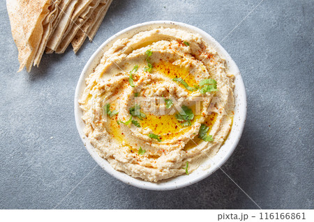 Bowl of creamy hummus garnished with olive oil and herbs is served with pita bread on a wooden board. A small bowl of garnish is placed beside the bread on a slate surface Bowl of creamy hummus garnished with olive oil and herbs is served with pita bread on a wooden board. A small bowl of garnish is placed beside the bread on a slate surface 116166861