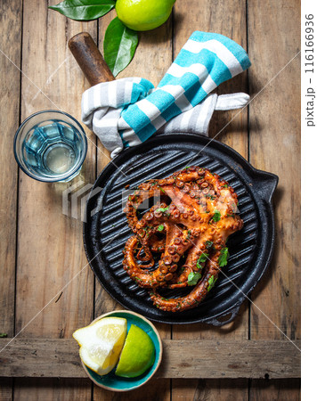 Grilled octopus on a black grill pan on a rustic wooden table, with limes, a blue-striped cloth Grilled octopus on a black grill pan on a rustic wooden table, with limes, a blue-striped cloth 116166936