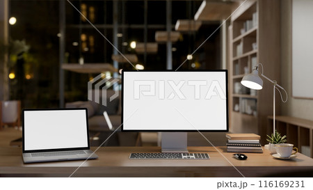 A modern office workspace at night features a laptop and computer mockup on a wooden table. 116169231