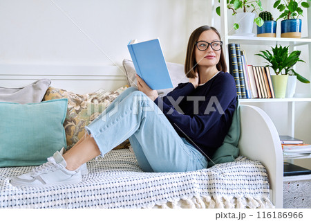 Young woman reading book, sitting on couch at home, engrossed in fiction 116186966