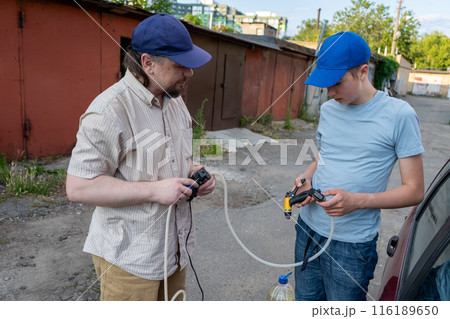 Dad and teenage son work together on the street near the garage. Outdoor activity. Family spend time. Helping and teach. Repairing a hose and pump for hand washing a car from a plastic bottle 116189650