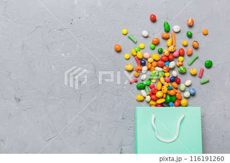 shopping paper gift bag in corner full of assorted traditional candies falling out on colored background with copy space. Happy Holidays sale concept 116191260