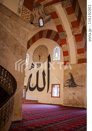 Interior of a mosque with large Arabic calligraphy on the walls and pillars with people are seen walking and praying on a red patterned carpet, in Edirne, Turkey. Interior of a mosque with large Arabic calligraphy on the walls and pillars with people are seen walking and praying on a red patterned carpet, in Edirne, Turkey. 116192801