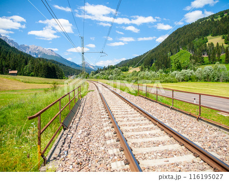 In the Dolomites, Italy, a set of train tracks runs through a scenic valley surrounded by green fields, forests, and snow-capped mountains under a bright blue sky. 116195027