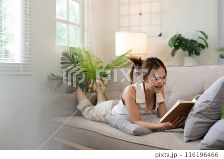 Smiling Asian woman reading book on couch in cozy living room. Concept of relaxation and comfort Smiling Asian woman reading book on couch in cozy living room. Concept of relaxation and comfort 116196466